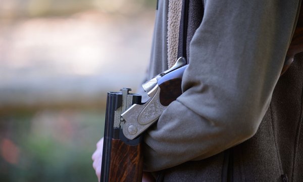 Eine Flinte liegt geöffnet im Arm einer Person, die eine dunkelgrüne Jacke trägt. Die Umgebung ist verschwommen und sieht nach Natur aus.