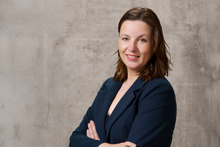 Eine Frau trägt einen dunklen Blazer und lächelt mit verschränkten Armen, stehend vor einer neutral grauen Wand.