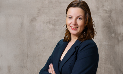 Eine Frau trägt einen dunklen Blazer und lächelt mit verschränkten Armen, stehend vor einer neutral grauen Wand.