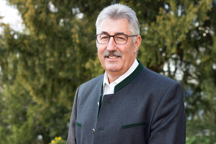 Ein Mann mit Brille und grauem Haar trägt einen traditionellen Janker, lächelt in die Kamera und steht vor einem Hintergrund aus grünen Bäumen.