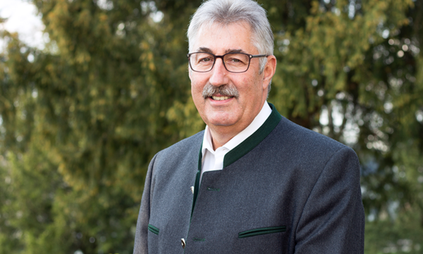 Ein Mann mit Brille und grauem Haar trägt einen traditionellen Janker, lächelt in die Kamera und steht vor einem Hintergrund aus grünen Bäumen.