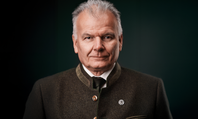 Ein älterer Mann trägt einen grauen Trachtenanzug und schaut ruhig in die Kamera. Er befindet sich vor einem dunklen, einfarbigen Hintergrund.