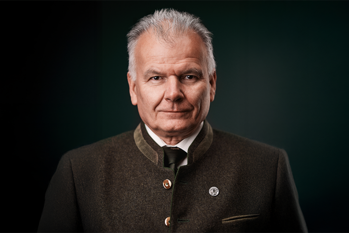 Ein älterer Mann trägt einen grauen Trachtenanzug und schaut ruhig in die Kamera. Er befindet sich vor einem dunklen, einfarbigen Hintergrund.