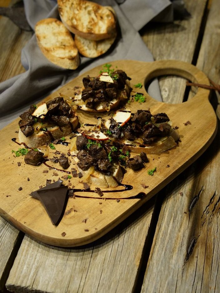 Geröstete Brotscheiben mit Fleischstückchen, Apfelscheiben und Kräutern, angerichtet auf einem Holzbrett. Daneben Schokoladenstücke, alles auf einem Holztisch mit grauer Serviette im Hintergrund.