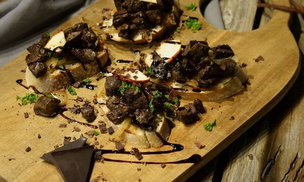 Geröstete Brotscheiben mit Fleischstückchen, Apfelscheiben und Kräutern, angerichtet auf einem Holzbrett. Daneben Schokoladenstücke, alles auf einem Holztisch mit grauer Serviette im Hintergrund.